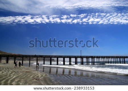 Ocean Pier. A pier goes into the ocean. People are strolling in the distance. A white cloud mirrors the pier in the sky.