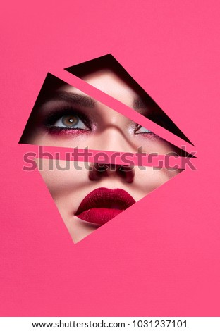 the face of a young beautiful girl with a bright make-up and with plump red lips peeks into a hole in pink paper.Fashion, beauty, cosmetics, make-up, beauty salon, make-up artist, sales, business.