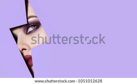 A girl with beautiful green bright eyes with blue shadows and bright purple lips looks into the hole of the colored paper.