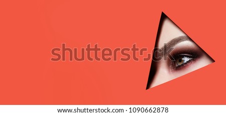 A girl with beautiful green bright eyes with brown shadows and expressive eyebrows looks into the hole of colored paper.