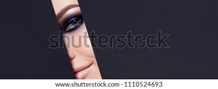 A girl with beautiful green bright eyes with blue shining shadows, beige lipstick and expressive eyebrows looks into the hole of colored paper.