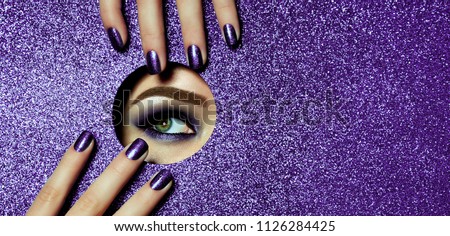 A girl with beautiful long fingers and lilac nails touches the edge of her open eye in a round hole of lilac shiny paper. A lilac eye shadow.