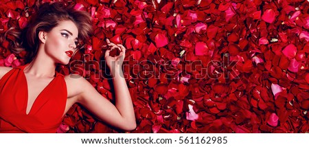 Valentine\'s Day. Loving girl. The girl in a red dress lying on the floor in the petals of red roses. Background of red rose petals. Red lipstick on the lips from the beautiful girl.