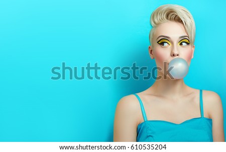 Fashionable girl with a stylish haircut inflates a chewing gum. The girl in the studio on a blue background. The girl\'s face with bright makeup and yellow with black shadows on the eyes.