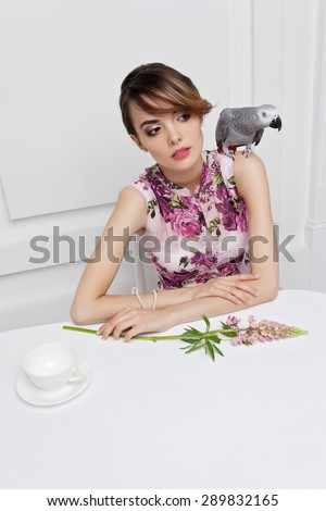 Fashionable attractive young woman in sitting in restaurant. With african grey parrot on shoulder and rose lupin flower on white table.
