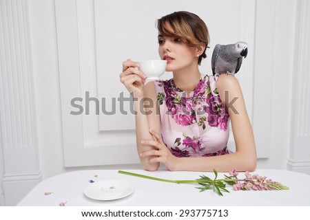 Beautiful Girl Drinking Tea or Coffee in Cafe.  With african grey parrot on shoulder, rose lupin flower on white table.