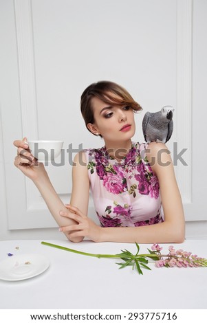 Beautiful Girl Drinking Tea or Coffee in Cafe.  With african grey parrot on shoulder, rose lupin flower on white table.