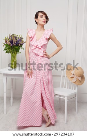Beautiful fashionable brunette standing near white table in long pink dress.