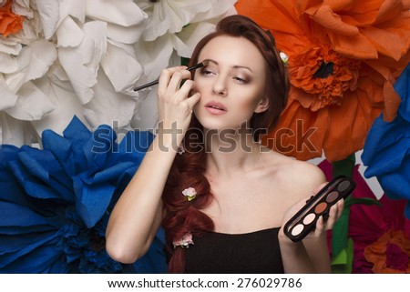 Close up beautiful women portrait with red hair,  doing beauty make up with brush eye shadow, long hair braided in a braid and small roses there and big colorful flowers on background