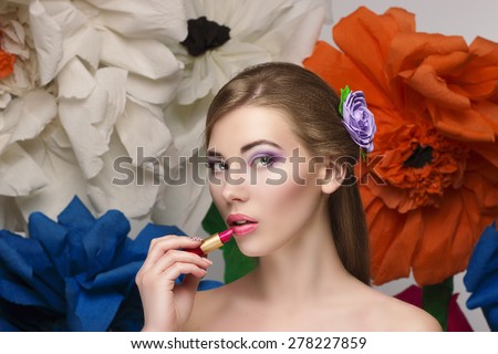 beautiful girl paints her lips with red lipstick and looks in the picture. Doing make up by herself with big summer flowers on bakcground