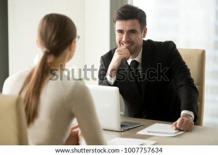 Handsome smiling male office worker in business suit sitting at desk with laptop in front of female colleague. Happy businessman talking with secretary. Positive first impression on job interview