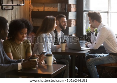 Diverse multiracial young people talking drinking coffee using devices in cozy coffeehouse, multi-ethnic african and caucasian millennials enjoy meeting sitting at coffeeshop tables together in cafe