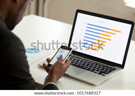 African american businessman using devices for business, black office worker holding smartphone working with laptop and mobile phone, corporate statistics apps and software, over the shoulder view