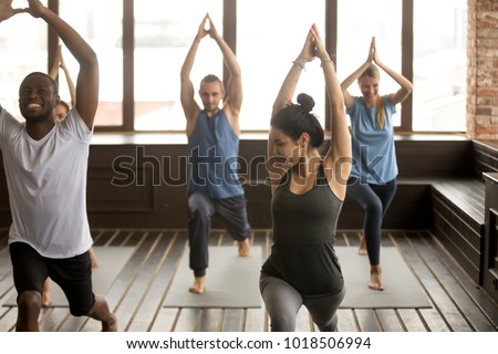 Group yoga in studio concept, diverse multiracial sporty fit people beginners practicing training with instructor teacher standing together trying to repeat warrior 1 pose, funny working out class
