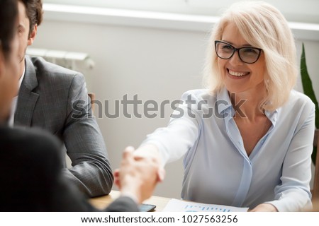Smiling attractive mature businesswoman handshaking businessman at meeting negotiation, happy hr senior executive woman shaking hand welcoming new hire partner, making deal satisfied with good result