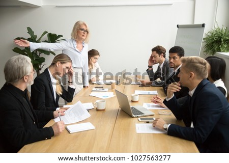 Angry senior woman boss firing unprofessional employee with hand gesture at diverse team meeting, dissatisfied aged female executive dismissing incompetent manager for bad work result in boardroom