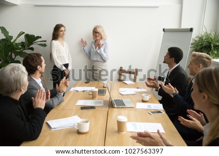 Senior woman boss introducing new female worker making compliment for work result while employees applauding at group meeting, team congratulating coworker with promotion, welcome recognition concept