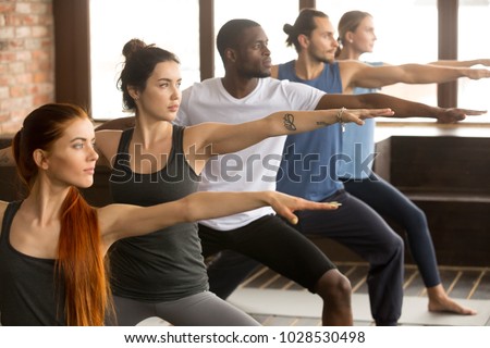 Group of young sporty afro american and caucasian people practicing yoga lesson, standing in Warrior two exercise, Virabhadrasana II pose, working out, indoor close up, studio background