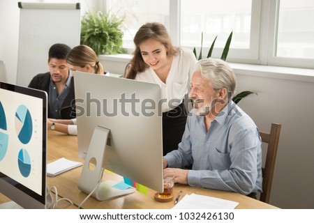 Smiling young manager helping senior worker with funny computer work in office, mentor teacher training happy older employee at workplace, colleagues of different age laughing looking at pc monitor