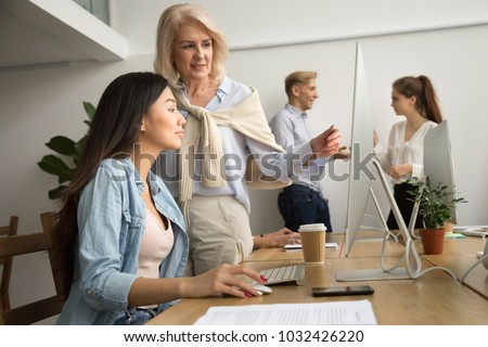 Aged female mentor training young asian intern explaining computer work, senior friendly executive teaching new employee looking at pc screen, older teacher talks to student helping with online task