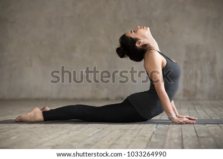 Young woman practicing yoga, doing Cobra exercise, Bhujangasana pose, working out, wearing sportswear, black pants and top, indoor full length, gray wall in yoga studio