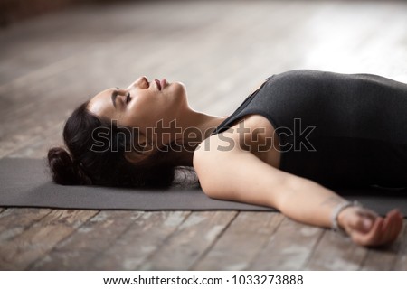 Young sporty woman practicing yoga, lying with eyes closed in Dead Body or Corpse pose, Savasana exercise, working out wearing sportswear top, resting after yoga, indoor close up photo, studio