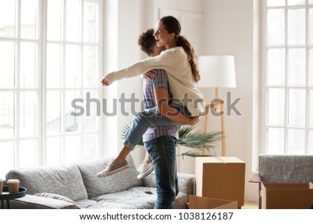 Excited couple celebrating moving day, man lifting embracing happy woman standing among boxes glad to move into new house, husband hugging delighted wife enjoying relocation starting living together