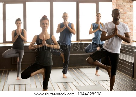 Group of young afro american and caucasian sporty people practicing yoga lesson standing in Vrksasana exercise, Tree pose with namaste gesture, working out, indoor, studio. Healthy lifestyle concept