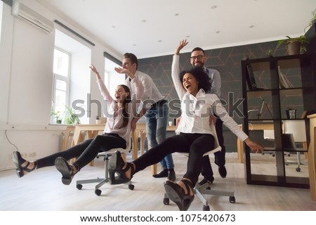 Millennial multiracial team people having fun riding on chairs in office room, excited diverse employees laughing enjoying funny activity at work break, creative friendly workers play game together