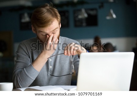 Young businessman taking off glasses feels eye strain tension tired of computer sitting at cafe table with laptop, millennial guy has bad sight vision problem massaging dry eyes after long laptop use