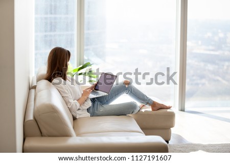 Relaxed woman using laptop in luxury home living room with big window, enjoying working, internet shopping, checking social network, reading news or communicating online with computer sitting on sofa