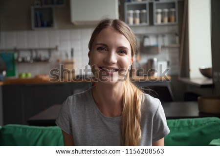 Smiling millennial woman sitting on kitchen sofa talking by videocall dating online looking at camera, video blogger vlogger recording vlog at home, lifestyle vlogging concept, head shot portrait