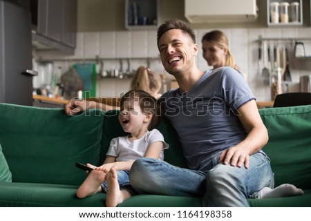Happy dad and kid son holding remote control laughing at funny humor comedy film or tv show sitting on sofa at home, smiling father having fun with child boy watching television together on weekend