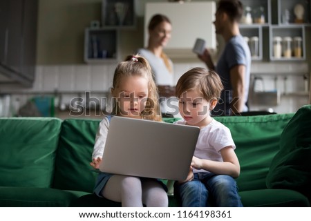 Cute children sister and brother watching online cartoons on laptop while parents talking in the kitchen, curious siblings boy girl using pc at home, kids computer addiction parental control concept