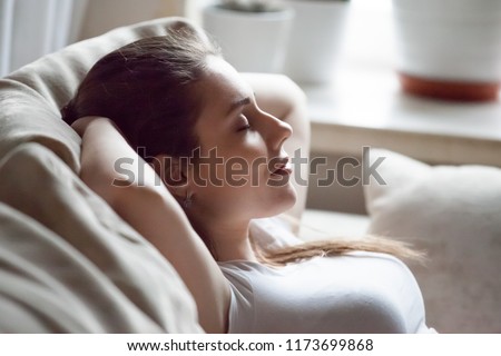 Calm pretty millennial woman lying on cozy sofa relaxing on weekend, peaceful female rest with eyes closed hands over head on couch, happy girl taking nap or chilling at home, sleep tired after work