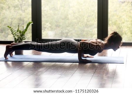 Young woman practicing yoga, doing four limbed staff, Push ups or press ups exercise, chaturanga dandasana pose, working out, wearing sportswear, grey pants and top, indoor full length, yoga studio