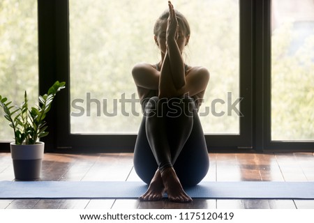 Young sporty attractive woman practicing yoga, doing Garudasana arms exercise, Eagle pose, working out, wearing sportswear, grey pants and top, indoor full length, yoga studio