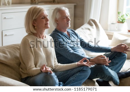 Calm senior middle aged couple practicing yoga together sitting in lotus pose on sofa, mindful peaceful mature man and woman meditating relaxing in living room at home, old people healthy lifestyle
