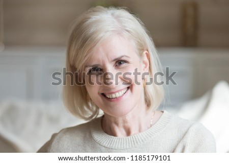 Happy middle aged elderly grey haired woman looking at camera posing at home indoors, positive satisfied single mature senior retired lady with wide toothy smile headshot portrait, natural old beauty