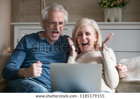 Excited senior middle aged old couple watching celebrating amazing victory winning online auction bid or bet together, elderly mature man and woman motivated by good news looking at laptop screaming