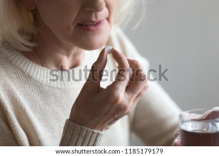 Mature senior middle aged woman holding pill and glass of water taking painkiller to relieve pain, medicine supplements vitamins, antibiotic medication, meds for old person concept, close up view