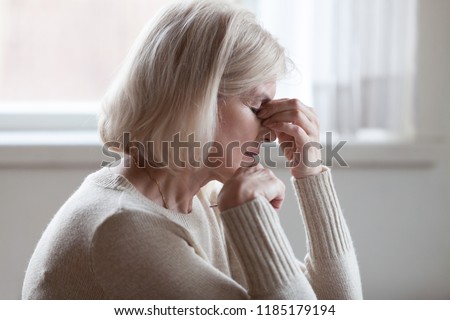 Fatigued upset middle aged older woman massaging nose bridge feeling eye strain or headache trying to relieve pain, sad senior mature lady exhausted depressed weary dizzy tired thinking of problems
