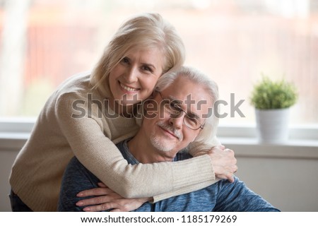 Smiling caring middle aged wife embracing senior husband at home, happy old woman hugging loving mature man looking at camera, retired elderly family married couple dating bonding headshot portrait