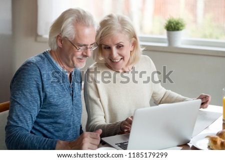 Happy old family couple talking using laptop having breakfast together, surprised excited senior woman looking at computer screen showing smiling middle aged husband online shopping sale on web site