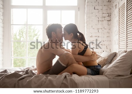 Excited couple kiss sitting in bed at hotel room. Young naked shirtless man embrace touch attractive girl in lingerie with love and desire. Romantic passionate relations, seduction foreplay concept