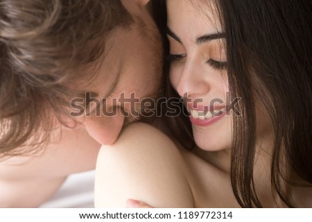 Close up portrait young attractive happy couple in love in bed. male gently with tender kisses shoulder of beloved naked smiling female. Passionate romantic relations and having sex concept
