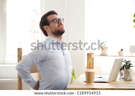 Tired millennial businessman suffering from backache feeling badly, sitting at office desk indoors. Have a back pain, sitting at the computer for a long time, sedentary work and bad posture concept