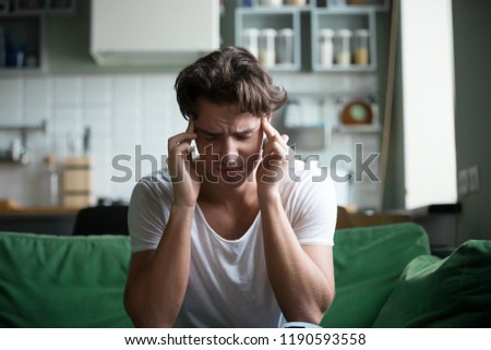 Young attractive stressed overworked person sitting on sofa with closed eyes touch head, having head ache or high blood pressure at home. Health problems or hangover after yesterdays hangouts, party