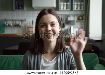 Head shot portrait of cheerful young woman sitting on sofa at home. Girl in casual clothes have video call using computer, wave her hand greeting or say goodbye to friend and smiling looking at camera