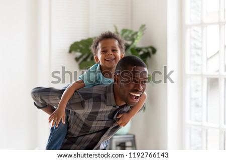 African black family spend time together have a fun standing in living room at home. Laughing father holding on back piggyback smiling cute little preschool toddler son Happy weekend free time concept
